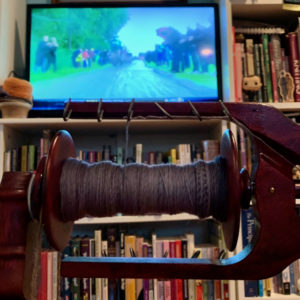Gray yarn on spinning wheel bobbin in foreground, television screen showing a bike race in the background