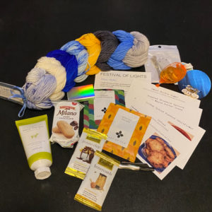 A set of mini-skeins of yarn in blue, gray, gold, and cream, several recipe cards, two packets of tea, chocolate gelt, stitch markers, cookies, and a few other treats.