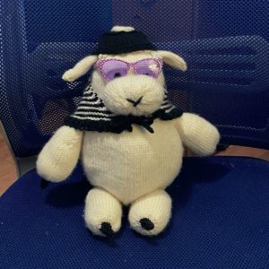 A knitted white sheep wearing purple-tinted glasses, a black hat, and a black and white shawl, sitting in a blue office chair